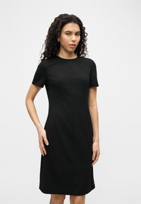 Γυναίκα με σγουρά μαλλιά φορόντας εφαρμοστό μαύρο φόρεμα με κοντά μανίκια, όρθια με το ένα χέρι στη μέση της μπροστά σε απλό φόντο.