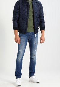 Granatowa pikowana kurtka bomberka na zielonym polo, w parze z przetartymi niebieskimi dżinsami i białymi trampkami. Teksturowany materiał, dopasowany fason.