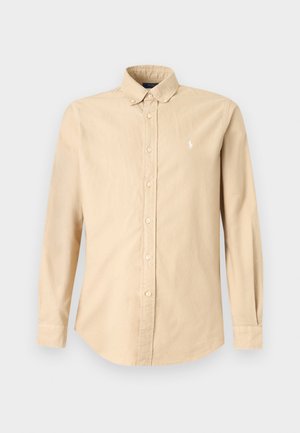 Mand iført beige skjorte med knapper, lyse lilla bukser, hvide sneakers og grå baseballkasket, stående mod en ensfarvet hvid baggrund.