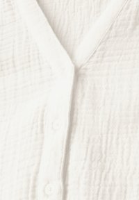 Hvid knap-op skjorte lavet af tekstureret stof, med en V-halsudskæring og små runde knapper foran til lukning.