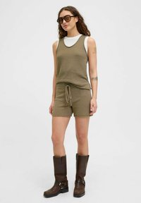 Pletena maslinasta majica bez rukava s odgovarajućim kratkim hlačama s vezicom u struku. Model nosi tamnosmeđe čizme do koljena i oversized sunčane naočale.