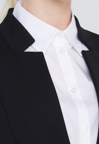 Blazer nero con collo a scialle, abbinato a una camicia bianca con bottoni trasparenti. Il tessuto ha una texture liscia.