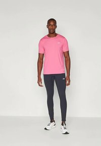 Bărbat îmbrăcat într-o cămașă roz cu mânecă scurtă, pantaloni sport negri bine adaptați și adidași albi, stând pe un fundal simplu.