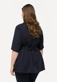 Blusa navy con maniche corte, caratterizzata da pois bianchi, vita arricciata con un fiocco e un tessuto testurizzato.