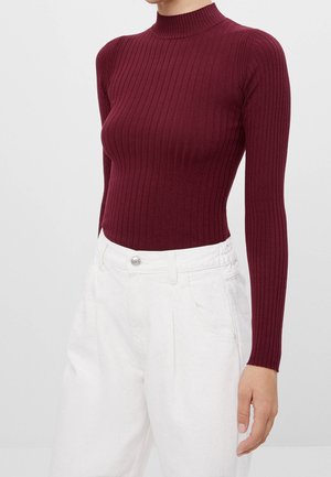 Burgunderfarbener gerippter Rollkragenpullover mit langen Ärmeln, taillierter Passform und hohem Kragen, kombiniert mit weißen hochtaillierten Hosen.