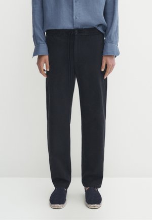 Mænds afslappede sorte bindebåndsbukser brugt sammen med marineblå slip-on sko og en blå skjorte med knapper, stående mod en ensfarvet hvid baggrund.