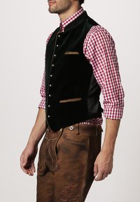 Dunkelgrüne Samtweste mit metallicfarbenen Knöpfen und braunen Akzenten, getragen über einem rot-weiß karrierten Hemd, kombiniert mit braunen Hosen.