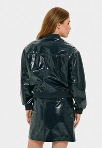 Giacca bomber lucida di un verde scuro con polsini e orlo a coste, caratterizzata da una texture liscia e lucida e una vestibilità su misura. I dettagli includono cuciture sui lati.