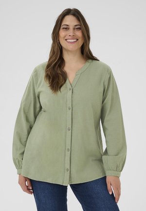 Usmievajúca sa žena v svetlozelenej košeli na gombíky s dlhými rukávmi a modrých rifliach, stojaca pred jednoliatym šedým pozadím.