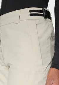 Beige pants with a textured surface, featuring a black adjustable strap at the waist, and side pockets for utility.