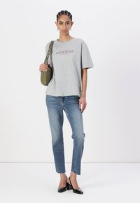 Šedé bavlněné tričko s logem "GUESS JEANS", v kombinaci s světle modrými rovnými džíny a černými lakovanými podpatky. Drží zelenou kabelku přes rameno.