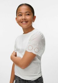 Chica sonriente con cabello oscuro, vistiendo una camiseta blanca con mangas de encaje, aretes de aro dorados y pantalones de mezclilla oscuros, frente a un fondo simple.
