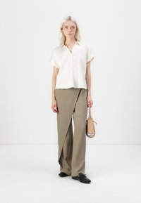 Weißes Kurzarm-Blusenshirt mit V-Ausschnitt und Knopfdetail, kombiniert mit khakifarbenen, weit geschnittenen Hosen. Tragetasche in Beige mit strukturiertem Tragegurt.