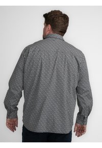 Man met bruin haar die een langwerpig shirt met patroon draagt, gezien van achteren tegen een eenvoudige witte achtergrond.
