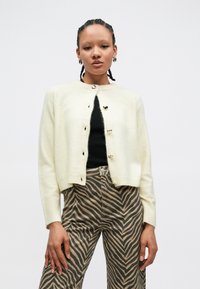 Cardigan en tricot crème avec un col rond et des boutons dorés en forme de cœur. Porté par-dessus un haut noir et associé à un pantalon marron à rayures zèbre.