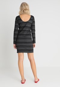 Kobieta z blond włosami stoi plecami, ubrana w czarną sukienkę z długim rękawem w zygzakowy wzór oraz czerwone płaskie buty na białym tle.