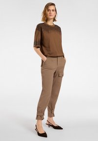 Brun bluse med pailletdetaljer, parret med lysebrune cargo bukser med sidelommer og knapdetaljer. Sorte slingback sko.
