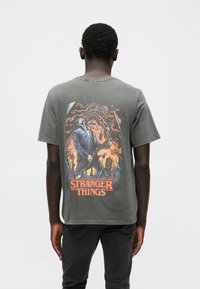 Gray cotton t-shirt featuring a vibrant graphic depicting a character with a guitar on the back, labeled "STRANGER THINGS" in orange.