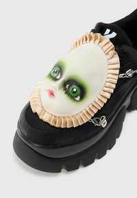 Scarpa piatta nera chunky con faccia di bambola decorativa che presenta occhi verdi, trucco scuro e bordo arricciato beige sulla parte superiore.