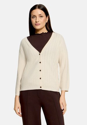 Woman wearing cream textured buttoned cardigan over dark brown high-neck top and matching pants, standing against plain background.