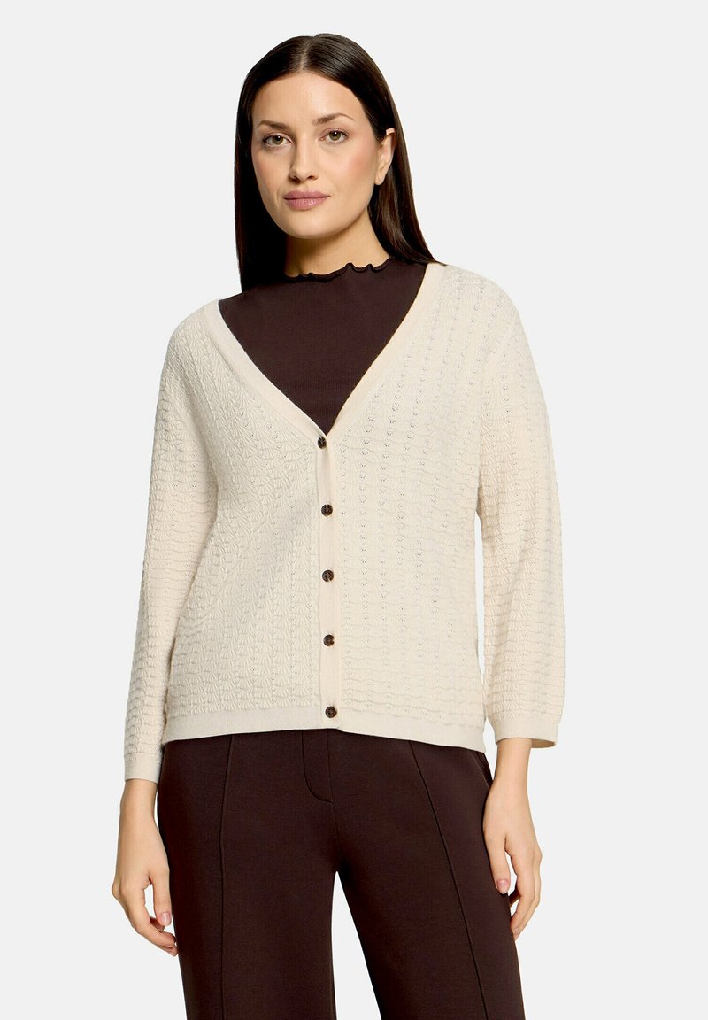 Femme portant un cardigan boutonné texturé crème sur un haut à col montant marron foncé et un pantalon assorti, debout devant un fond uni.