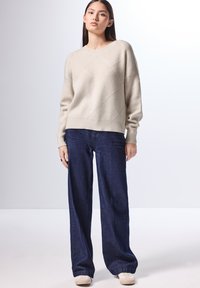 Lichtbeige gebreide trui met een gestructureerd ruitpatroon, gecombineerd met donkere blauwe wijde jeans en lichtgekleurde schoenen.