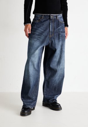 Jean en denim bleu à jambes larges avec une teinture délavée, présentant des poches frontales inclinées, des coutures visibles et un ourlet légèrement effiloché. Bottes noires portées.