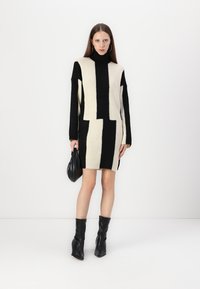 Adīta kleita ar augstu apkakli, kurai ir drosmīgs melnbalts ģeometrisks raksts. Valkāta ar melniem ceļgala augstiem zābakiem un mazu somiņu.