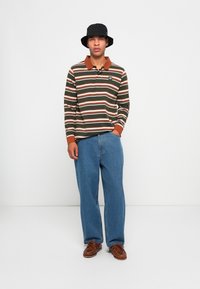 Langsleeve gestreepte polo in donkergroen met witte en roestkleurige strepen, voorzien van een roestkleurige kraag en logo patch, gecombineerd met losse blauwe jeans en bruine schoenen.