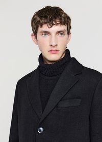 Schwarzer Wollmantel mit einem einzelnen Knopf und einer Tasche, darüber ein schwarzer, gestrickter Rollkragenpullover. Glatte Textur, maßgeschneidertes Design, moderne Passform.