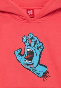 Moletom vermelho com capucho apresentando uma ilustração de uma mão azul com a boca aberta e a língua de fora, exibindo linhas pretas para detalhes e um logotipo "Santa Cruz".