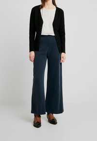 Cardigan noir avec des boutons superposé sur un top texturé blanc, associé à un pantalon large navy et des chaussures noires ornées.