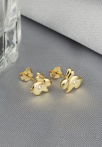 Boucles d'oreilles en forme de lapin en or avec une finition polie et des accents de pierres précieuses transparentes, sur un fond de tissu gris texturé.