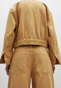 Beige gestrickte Jacke mit elastischem Saum und weiten Ärmeln, kombiniert mit hochtaillierten Hosen mit zwei Gesäßtaschen. Weicher Stoff mit angenehmer Textur.