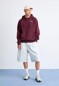 Maroon hoodie with a front pocket and logo, paired with light blue shorts, white ankle socks, and light sneakers with black soles.
