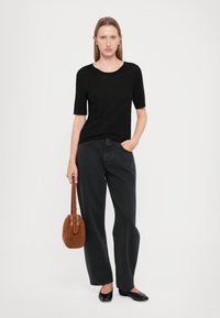 Musthave musta lühikeste varrukatega top ja laiad mustad teksad, hoides käes pruuni nahast kotti ja jalas mustad lamedamad kingad. Neutraalne taust.