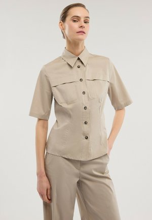 Frau mit zurückgebundenem Haar, trägt ein beigefarbenes, kurzärmeliges Hemd mit Knöpfen und passende beige Hose, steht mit einer Hand in der Tasche.
