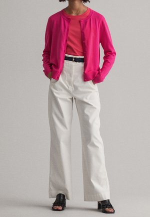 Person trägt eine leuchtend pinke Strickjacke über einem roten Oberteil, weiße hoch taillierte Hose mit schwarzem Gürtel und schwarze, offene, mit Absatz versehene Schuhe.