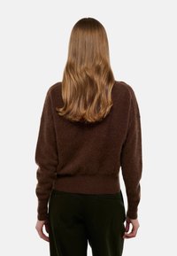 Pull marron, fluffy avec une coupe décontractée, poignets et ourlet côtelés. Associé à un pantalon vert foncé, vu de dos.