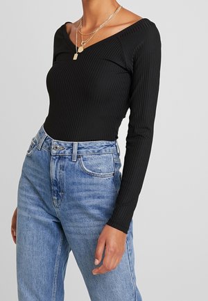 Frau trägt ein schwarzes, geripptes Langarmtop mit off-shoulder Ausschnitt und geschichteten goldenen Halsketten, kombiniert mit hoch taillierten blauen Jeans.