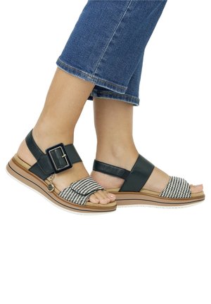 Des pieds portant des sandales noires et blanches à bout ouvert avec des brides à la cheville et une grande boucle, assorties à un jean en denim bleu.