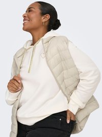 Beige vadderad väst med quiltad design över en krämfärgad huvtröja med en subtil hjärtlogotyp. Modellen bär mörka jeans och små ringörhängen.