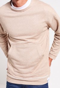 Béžová mikina se strukturovaným vzorem, kulatým výstřihem a přední kapsou. Dokončeno kontrastním šitím a božní etiketou.