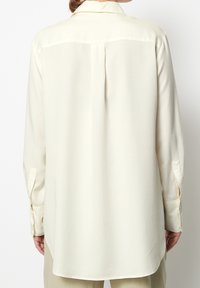 Lichtbeige blouse met lange mouwen en een klassieke kraag, gladde textuur en een ronde zoom. Bevat een schouderstukdetail en knoopmanchetten.
