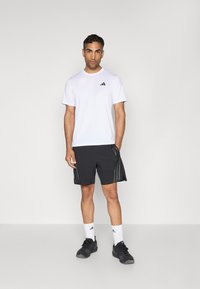 Weißes Kurzarm-T-Shirt mit schwarzem Adidas-Logo, kombiniert mit schwarzen Shorts mit seitlichen Streifen, weißen Socken und schwarzen Sportschuhen.