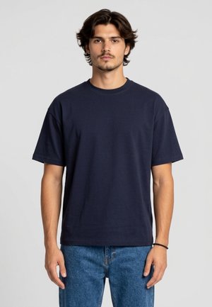 Giovane uomo con capelli scuri che indossa una maglietta blu navy semplice e jeans blu, in piedi davanti a uno sfondo grigio chiaro.