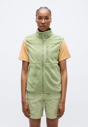 Gilet verde in pile con collo alto, zip frontale e tasca sul petto, indossato sopra una maglietta beige a maniche corte e pantaloncini verdi.