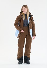 Braune Schneejacke mit hellblauen Akzenten und schwarzem Innenfutter, kombiniert mit braunen Schneehosen. Verfügt über Reißverschlusstaschen und verstellbare Ärmelabschlüsse.