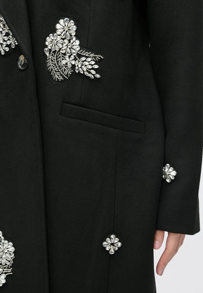 Črn plašč z bogatimi cvetličnimi kristalnimi okrasitvami na prsih in rokavih, z elegantnim, prirejenim oblikovanjem in stransko žepom.