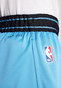 Modri košarkarski šortsi z mrežasto teksturo, črn elastičen pas s svetlo modrimi črtami, na levi strani pa ima logo NBA.